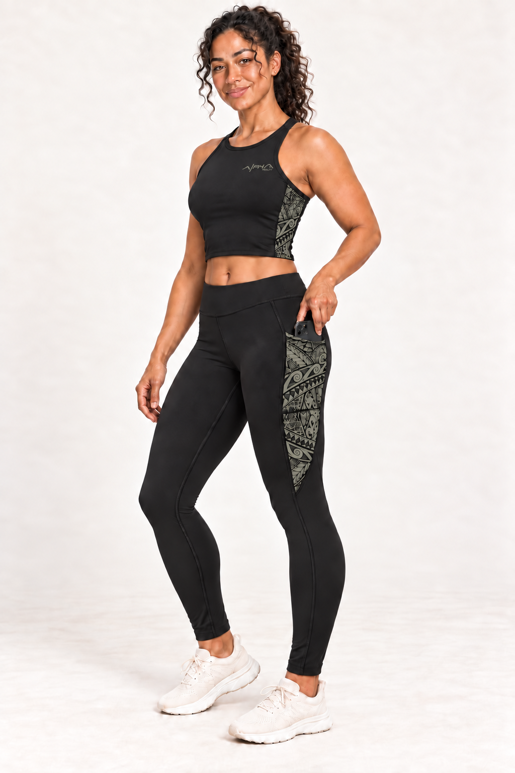 Model wearing black Alpha Maui Trail Runner leggings with muted olive tribal artwork and matching crop tank, placing a phone in the side pocket.