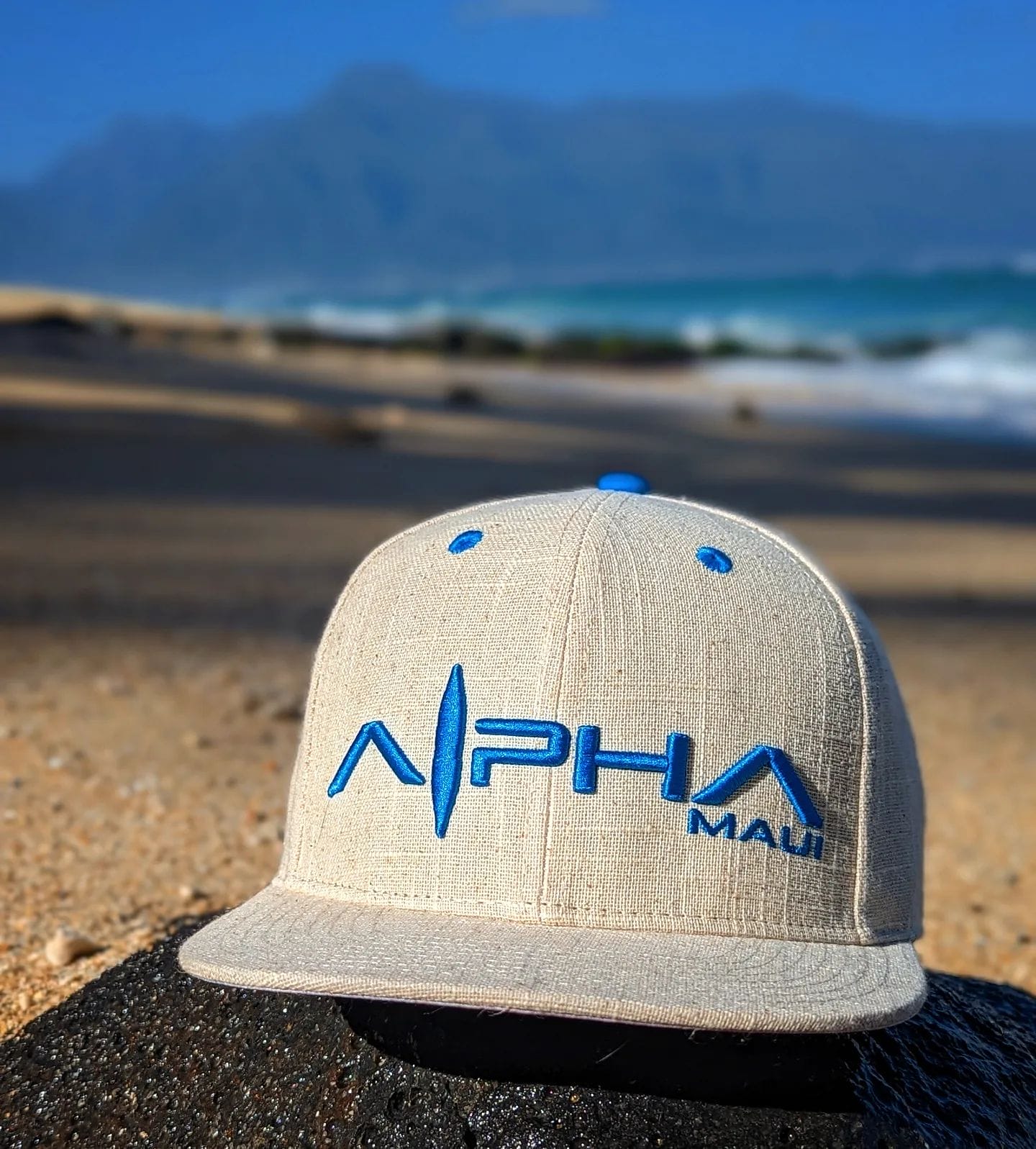 Close-up of a beige baseball cap with blue text that reads "Alpha Maui." The cap, a perfect piece of beach apparel, is resting on a rock with sandy shores and ocean waves in the blurred background. The clear sky and distant mountains complete this Hawaii scene infused with Ohana spirit.