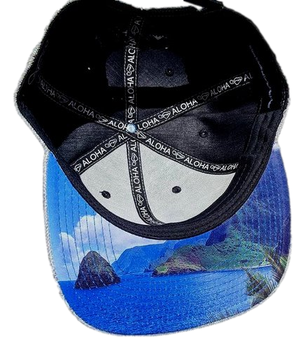 The Tinsman cap features a black and white design with the word "Aloha" repeated on its black interior straps. The underside of the brim displays a vibrant ocean scene complete with rocky cliffs and a clear blue sky, making it perfect beach apparel for anyone embracing the Alpha Maui spirit.