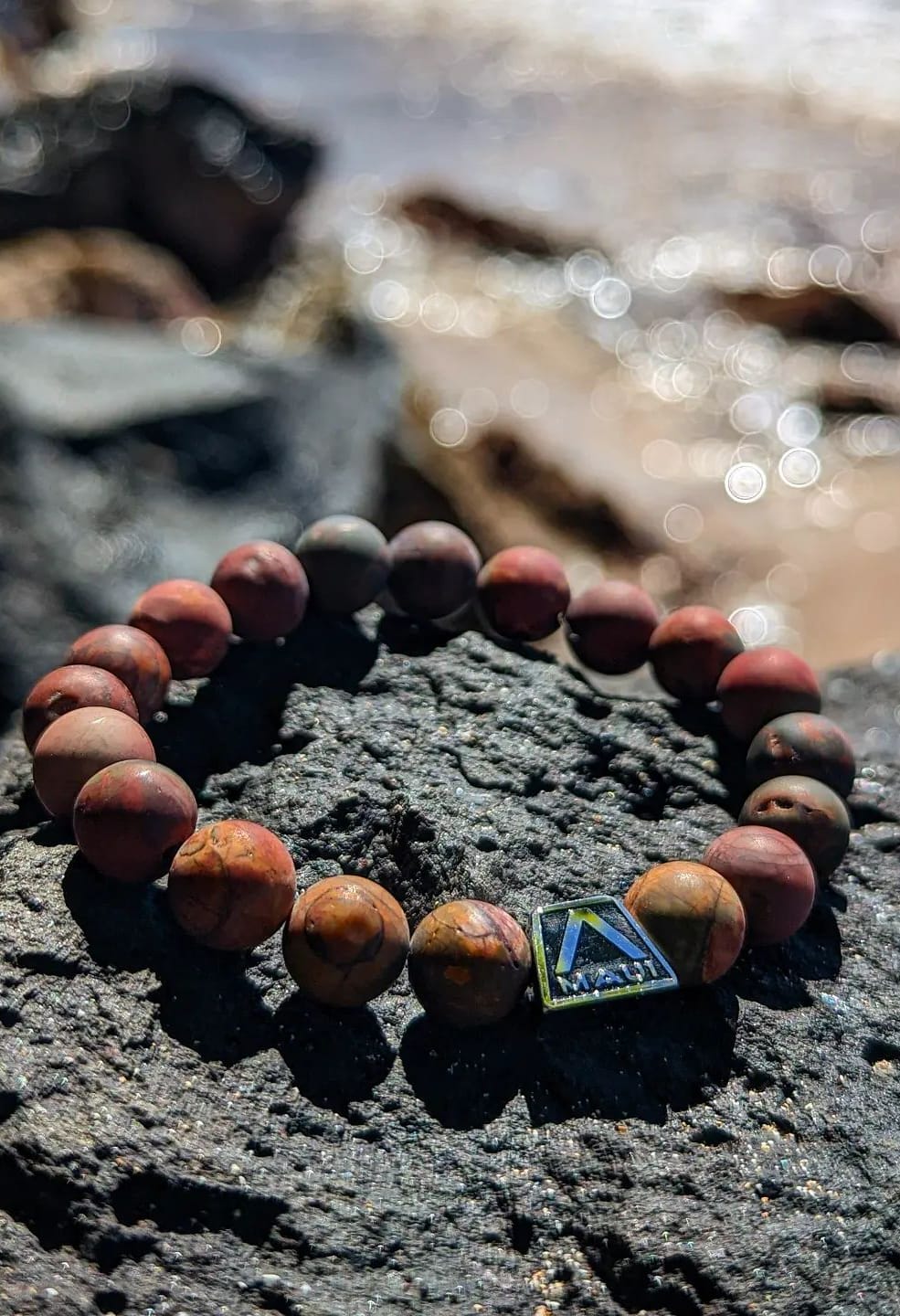 A beaded bracelet with natural stone beads and a small, metallic pyramid-shaped charm is displayed on a dark rock surface. The background shows a blurry, sunlit beach with gentle waves, making it the perfect piece for your Alpha Maui beach apparel collection.