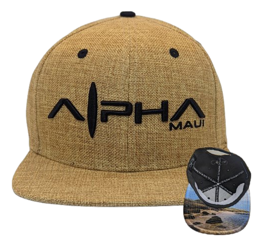 The Hemped Out baseball cap features a tan color with black embroidered text reading "Alpha Maui" on the front. The underside of the brim showcases an illustrated coastal landscape with rocks, ocean, and sky, perfectly capturing the spirit of Hawaii.