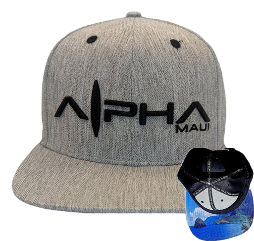 The Tinsman is a gray snapback hat featuring black embroidery that spells "Alpha Maui" on the front. Two black ventilation holes are visible at the top. The interior of the hat, shown in a small overlay, displays a blue ocean print, making it a perfect addition to your beach apparel collection and reminiscent of Hawaii's Ohana spirit.
