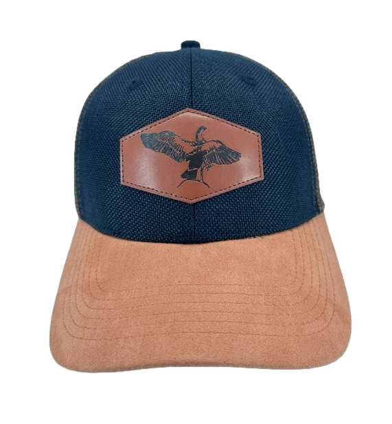 The Alpha Maui NeNe, a dark blue baseball cap with a light brown suede brim, is perfect for beach apparel. It features a brown leather patch on the front with a black silhouette of a goose in flight. The simple and outdoorsy design embodies the Ohana spirit.