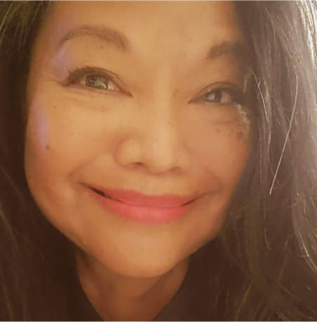 A person with long dark hair smiles warmly at the camera, with soft lighting highlighting their features. They are wearing makeup, including pink lipstick, and have a gentle, cheerful expression.