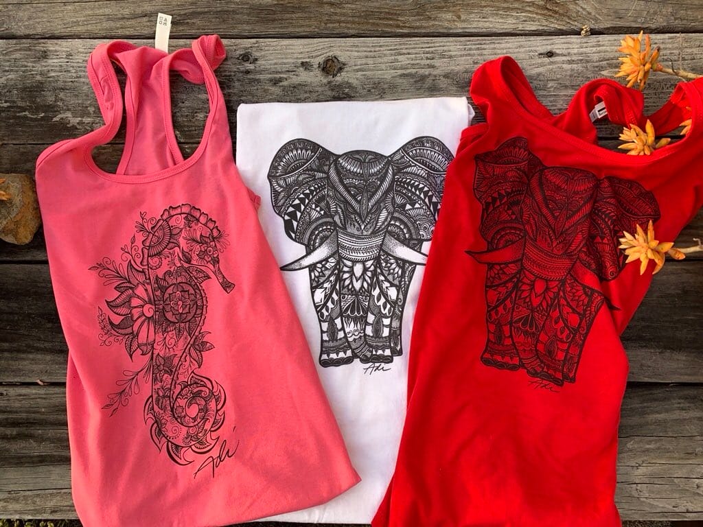 Three tank tops with intricate black elephant designs: a pink one with a side profile, a white one with a front view, and a red one with a detailed front view. They are displayed on a wooden surface with orange flowers.