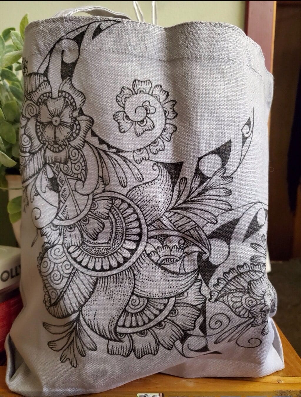 A beige fabric tote bag with an intricate black floral and abstract design is placed on a wooden surface. In the background, there is a potted plant and a stack of books.