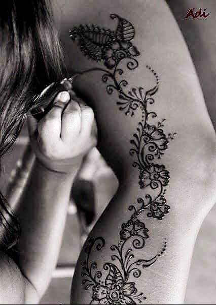 A person applying intricate floral and paisley henna designs on someone's arm. The image is in black and white, highlighting the detailed artwork.