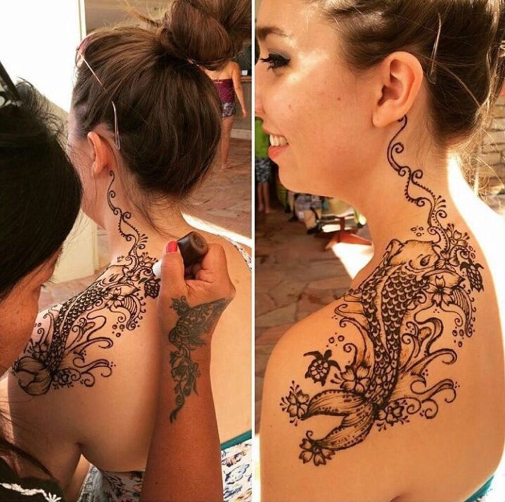 A woman is getting a henna tattoo on her shoulder and back. The design features an intricate koi fish pattern with floral elements. Another person is applying the tattoo with a henna cone. The setting appears to be outdoors.